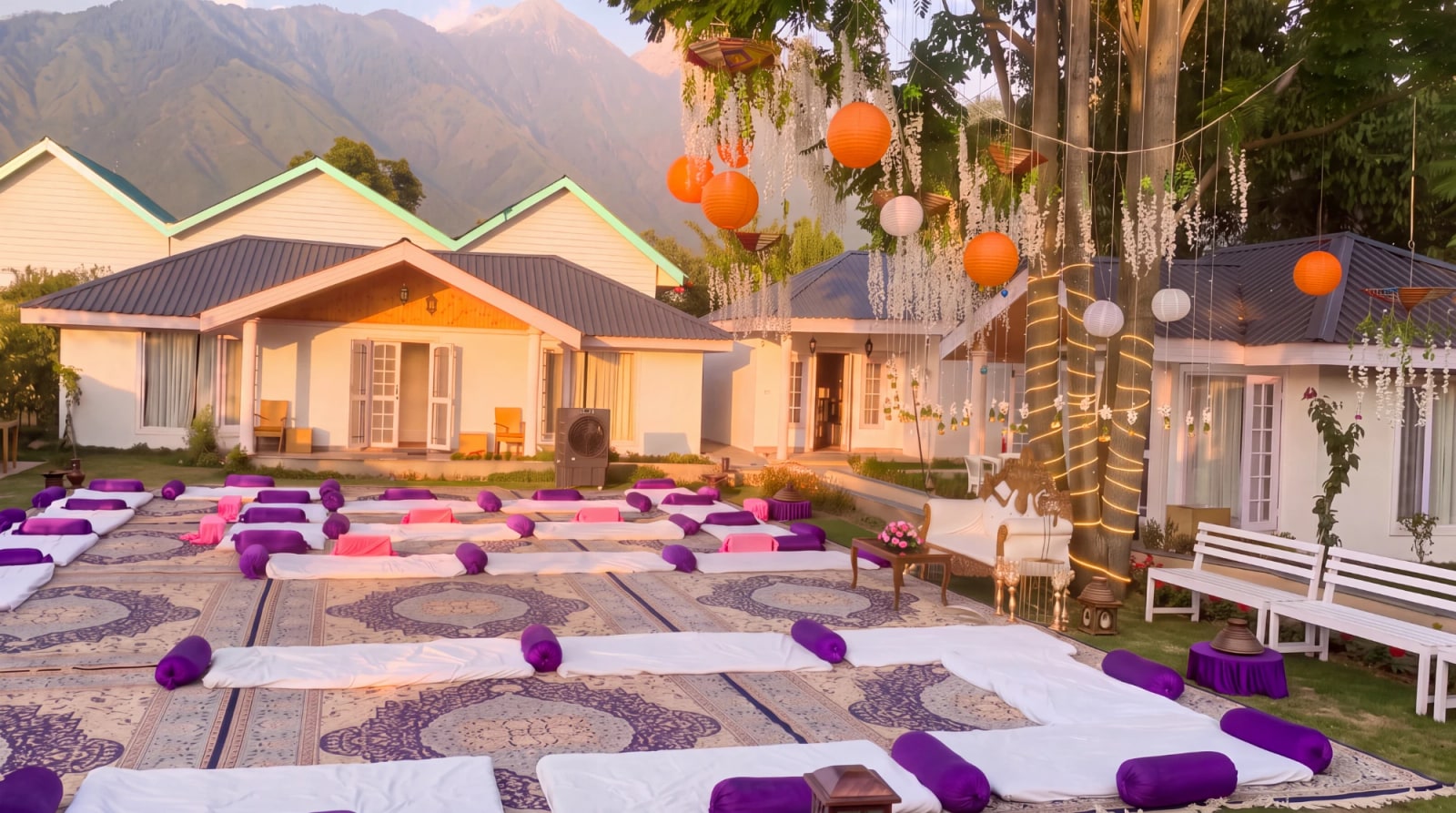 Floor seating laid out on carpets for a wazwan, with lanterns strung through the trees and the Zabarwan behind